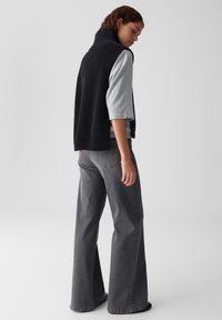 Frau trägt eine ärmellose schwarze Strickweste über einem hellgrauen kurzärmeligen Oberteil und hochtaillierten, weiten grauen Jeans, schaut nach unten und steht.