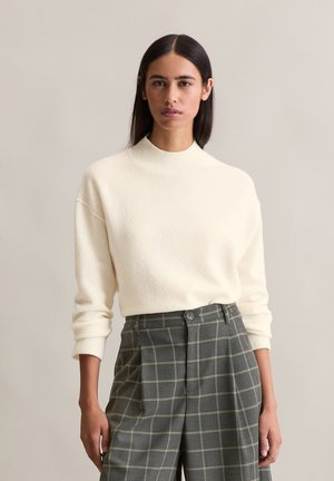 Cream-colored, knitted sweater with a high ribbed neckline and dropped shoulders, paired with gray checkered trousers featuring yellow lines.