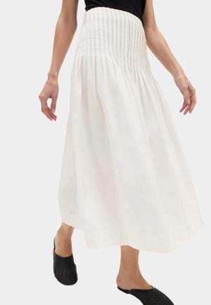 Frau trägt einen weißen, plissierten Midi-Rock mit hoher Taille, kombiniert mit schwarzen gewebten Slip-On-Schuhen und einem schwarzen ärmellosen Oberteil.