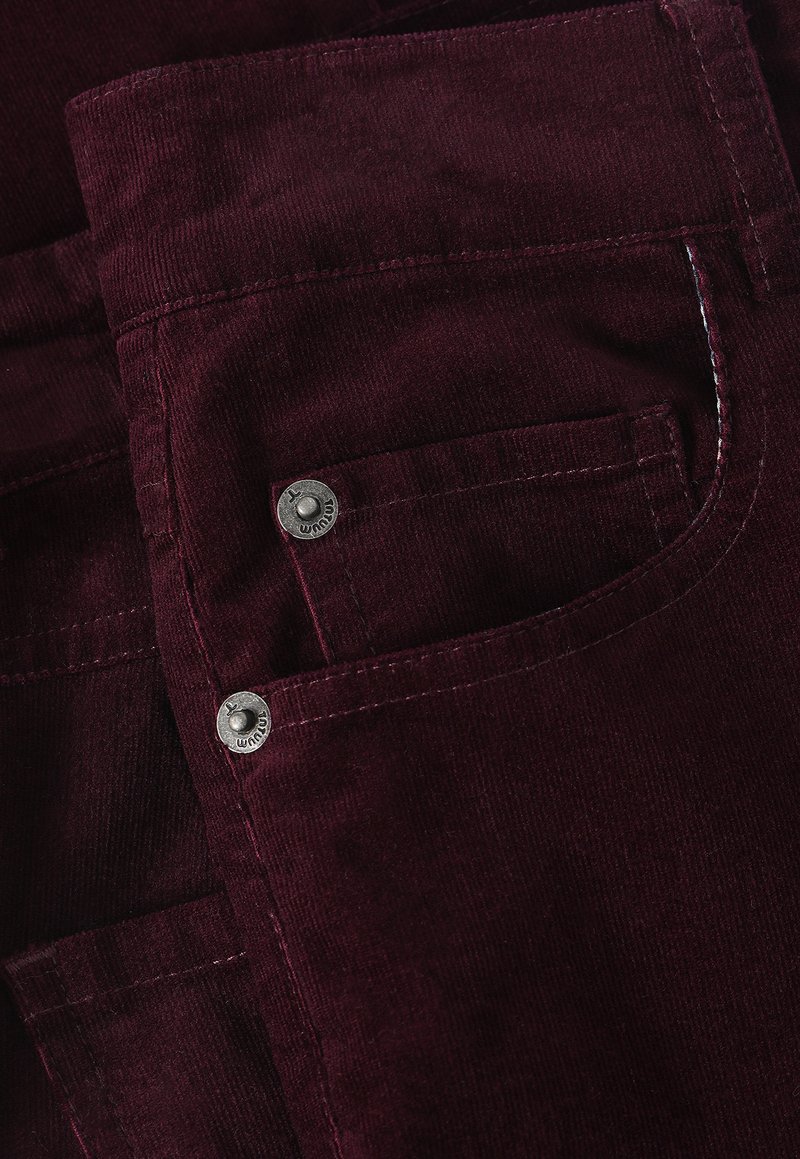 Bordowe sztruksowe spodnie z dwoma srebrnymi metalowymi guzikami, podkreślające teksturowany materiał i staranne przeszycia. Widoczny detal kieszeni.