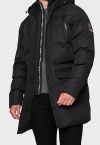 Giubbotto invernale imbottito nero con cappuccio, tasche con zip e chiusure a pressione. Presenta una toppa con logo e un dettaglio della zip interna in contrasto.