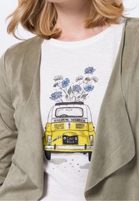 Olivegrøn ruskindsjakke over en hvid t-shirt med en gul vintagebil med blå blomster og "CIAO BELLA" på nummerpladen.