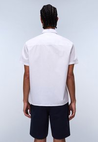 Kurzärmliges weißes Hemd mit geradem Schnitt, glatter Textur und klassischem Kragen, kombiniert mit dunklen Shorts. Rückansicht.