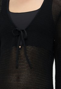 Black crochet top featuring a deep V-neck, tied front closure, and a sheer texture, paired with a solid black undershirt.