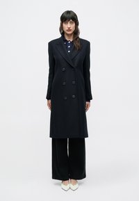 Zwarte dubbelknoops jas met grote knopen, een getailleerde pasvorm en een strakke silhouet. Gecombineerd met zwarte broek met brede pijpen en puntige witte schoenen.