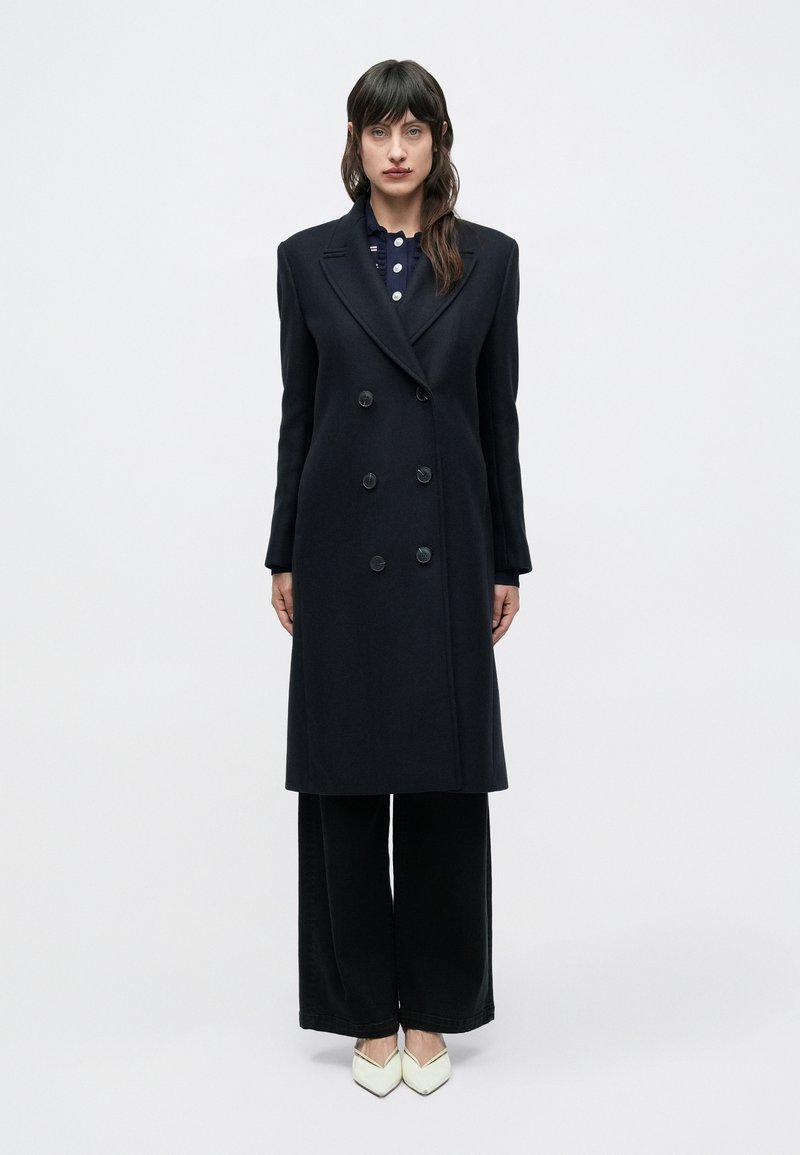 Zwarte dubbelknoops jas met grote knopen, een getailleerde pasvorm en een strakke silhouet. Gecombineerd met zwarte broek met brede pijpen en puntige witte schoenen.