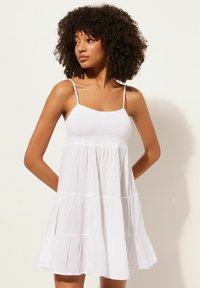 Weißes Baumwollkleid mit Spaghettiträgern, gerafftem Oberteil und gestuftem Rock. Leichter, fließender Stoff und subtile Textur unterstreichen das Design.