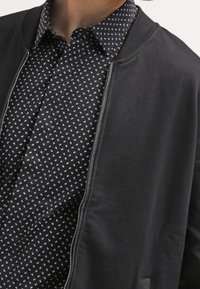 Giubbotto bomber nero con una texture liscia e zip, indossato sopra una maglietta nera con un motivo a quadrati bianchi.
