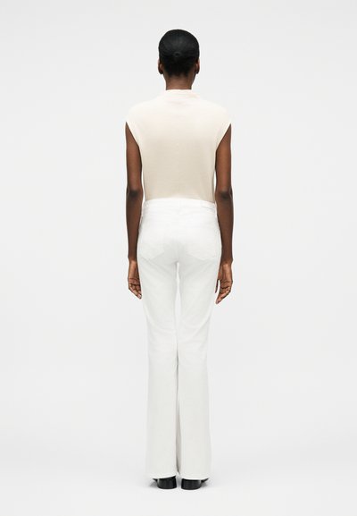 Γυναίκα με σκούρο χρώμα δέρματος και μαλλιά σε κότσο, που στέκεται με την πλάτη προς τα έξω, φοράει αμάνικο μπεζ τοπ, λευκό παντελόνι και μαύρα παπούτσια σε λευκό φόντο.