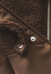Veste isolante marron en tissu moelleux et texturé avec une finition satinée lisse. Présente des boutons-pression et une fermeture éclair métallique sur le côté.