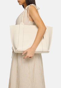 Grande borsa a tote di colore crema con design strutturato, materiale in ecopelle liscia e dettagli minimali. Modella che la tiene all'altezza della spalla.