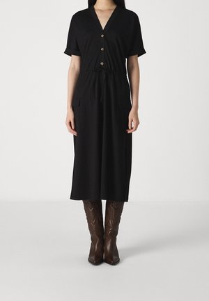 Vrouw draagt een zwart midi-jurk met knopen en een tailleceintuur, gecombineerd met kniehoge bruine leren laarzen, staand op een effen achtergrond.