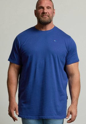 Brodaty mężczyzna z krótkimi włosami stoi i patrzy przed siebie, ma na sobie zwykły niebieski T-shirt i jasnoniebieskie dżinsy na neutralnym tle.