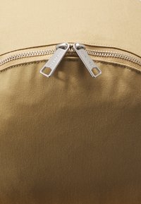 Beige stoffen tas met een gladde textuur, voorzien van dubbele zilveren ritsen en een gestikte naad. De kleur is uniform zonder patronen.