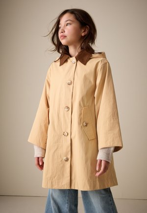 Joven vestido con un abrigo beige abotonado con cuello marrón y vaqueros azules, de pie frente a un fondo liso claro.
