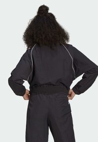 Giacca a vento nera con vita e polsini elasticizzati, piping bianco a contrasto lungo le spalle, tessuto leggero e vestibilità rilassata.