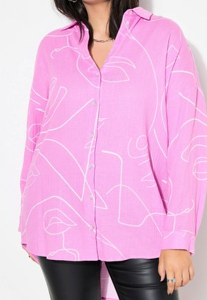 Frau trägt ein weites rosa Hemd mit abstraktem weißen Linien-Gesichtsprint und schwarze Lederhose, steht vor einem weißen Hintergrund.