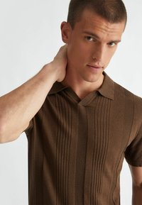 Polo di maglia marrone con motivi a coste verticali e texture. Presenta un colletto classico e maniche corte. Finitura del tessuto liscia.