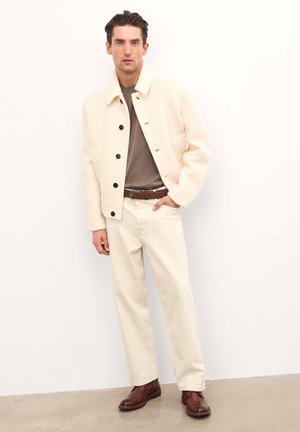 Chaqueta blanca de corte relajado con botones negros, camisa marrón, pantalones crema y zapatos marrones. Textura suave, diseño informal.