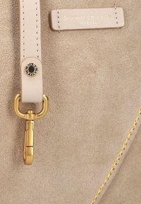 Beige suède handtas met een gouden sluiting, zachte textuur en contrasterende gele stiksels; gegraveerd logo op een glad leren label.