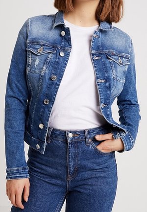 Woman wearing blue denim jacket over white t-shirt, paired with high-waisted blue jeans, hand in pocket, standing against plain background.