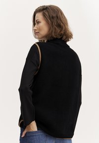 Gilet in pile nero con colletto alto, rifiniture brown a contrasto sulle spalle, abbinato a una maglietta a maniche lunghe e jeans di denim. Texture liscia.