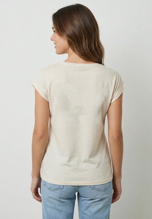 Donna con capelli castani lunghi che indossa una maglietta beige semplice a maniche corte e jeans azzurri, vista da dietro su uno sfondo uniforme.