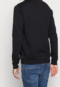 Pull en laine noir avec des manches longues, des poignets et un ourlet côtelés, texture lisse, et un petit détail décoratif près du bas gauche.