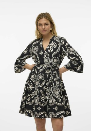 Robe noire avec des motifs floraux et de feuilles en crème. Dotée d'un décolleté en V, de manches longues évasées et d'un détail à nouer à la taille. Design de jupe évasée.