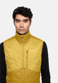 Gelbe isolierte Weste mit hohem Kragen und Reißverschluss. Mit Zickzackmustern und einem Logo auf der Brust. Schwarzes Langarmshirt darunter.