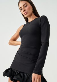 Black dress with asymmetrical long sleeve, fitted bodice, and ruffled hem. Smooth texture, minimalist design, and sleek silhouette.
