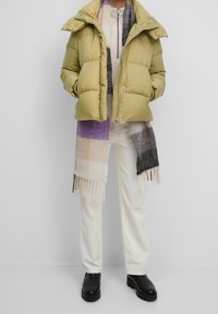 Oliwkowa kurtka puchowa z wysokim kołnierzem, zestawiona z wzorzystym szalikiem w kolorach fioletowym, beżowym i szarym. Białe spodnie kordowe i czarne botki.