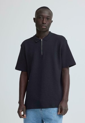 Mann iført en mørk marineblå kortermet polo med glidelåsdetalj, kombinert med lyseblå jeans, stående foran en enkel lys bakgrunn.