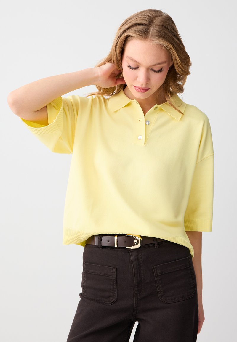 Femme aux cheveux blonds portant un polo jaune à manches courtes rentré dans un pantalon noir taille haute avec une ceinture noire, regardant vers le bas.