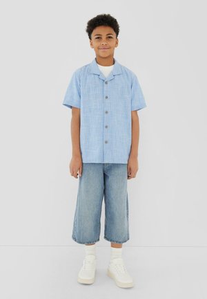 Chłopiec ubrany w jasnoniebieską koszulę z krótkim rękawem zapinaną na guziki, białą koszulkę pod spód, szerokie niebieskie spodnie dżinsowe do połowy łydki, białe skarpetki i białe trampki, stoi.