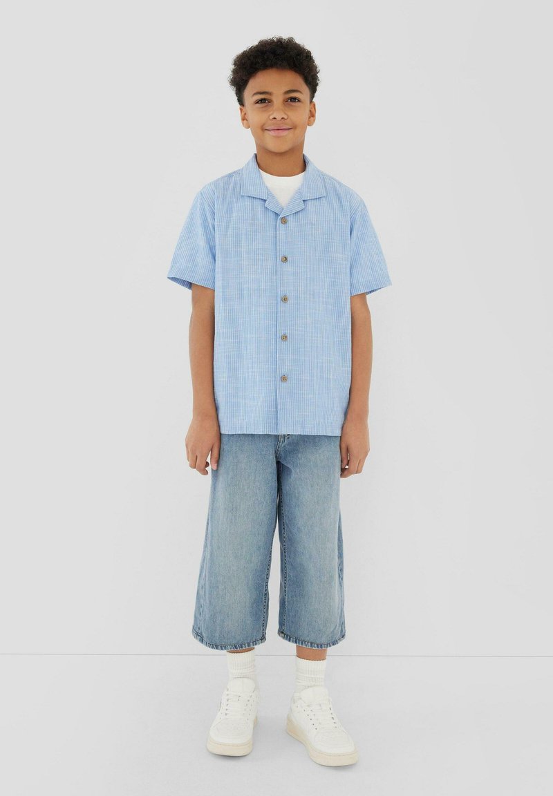 Garçon portant une chemise bleu clair à manches courtes avec des boutons, un t-shirt blanc en dessous, un pantalon large en jean bleu raccourci, des chaussettes blanches et des baskets blanches, debout.