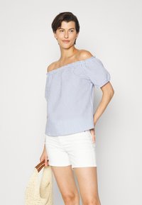 Top azul e branco às riscas, com ombros à mostra e mangas bufantes, de corte solto, combinado com calções brancos e uma bolsa de palha.