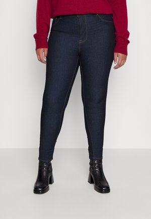 Osoba ubrana w obcisłe, ciemnoniebieskie jeansy, czarne botki na obcasie oraz czerwoną, długorękawową bluzę, stojąca na jednolitym tle.