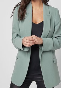 Donna che indossa un blazer verde salvia sopra una maglietta nera e pantaloni neri, con le mani dolcemente incrociate davanti.