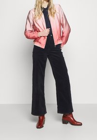 Lichtroze satijnen bomberjack met een verloopkleur, witgeribbelde mouwen met strepen, gecombineerd met marineblauwe wijde corduroy broek en rode enkellaarsjes.