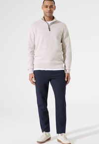 Suéter de pullover beige con media cremallera, sobre una camisa blanca, combinado con pantalones azul marino y zapatillas blancas con suelas marrones. Textura suave, diseño casual.