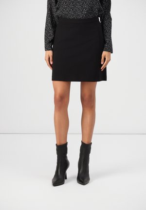 Femme portant une jupe noire longueur genoux, un haut noir à manches longues à motifs et des bottines noires à talons, debout sur un sol blanc uni.