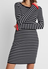 Blau-weiß gestreiftes Strickkleid mit langen Ärmeln, mit gerippter Struktur und rotem Akzent an den Ärmelbündchen. Figurbetonte Silhouette bis zur Kniehöhe.