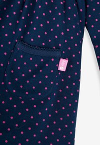 Material bleu marin cu buline roz, având o buzunar cu margini fluturate și o etichetă mică roz. Textură moale și design casual.