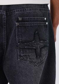 Jeans neri in denim con un motivo di cuciture nere strutturate sulla tasca posteriore. Presentano dettagli in metallo argentato e tessuto scuro sbiadito.