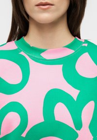 Žena u ružičastoj košulji s velikim apstraktnim zelenim oblicima, prikazana od vrata do gornjeg dijela prsa na neutralnoj pozadini.