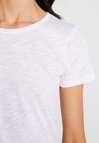 Bílé tričko s krátkým rukávem vyrobené z lehkého texturovaného materiálu, s kulatým výstřihem a mírně přiléhavým střihem. Žádné viditelné vzory nebo akcenty.