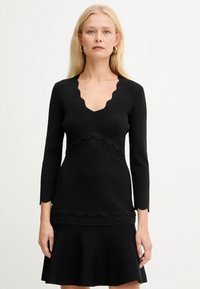Donna bionda che indossa un vestito nero a maniche lunghe con bordi a scallop sulla scollatura, in vita, sulle maniche e sull'orlo, su uno sfondo semplice.