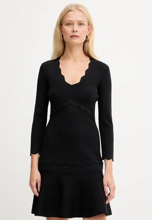 Donna bionda che indossa un vestito nero a maniche lunghe con bordi a scallop sulla scollatura, in vita, sulle maniche e sull'orlo, su uno sfondo semplice.