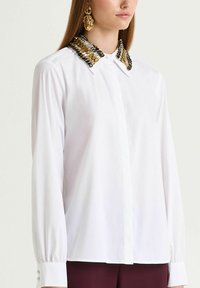 White shirt with a patterned collar featuring gold and black accents, long sleeves with buttoned cuffs, and a straight silhouette.